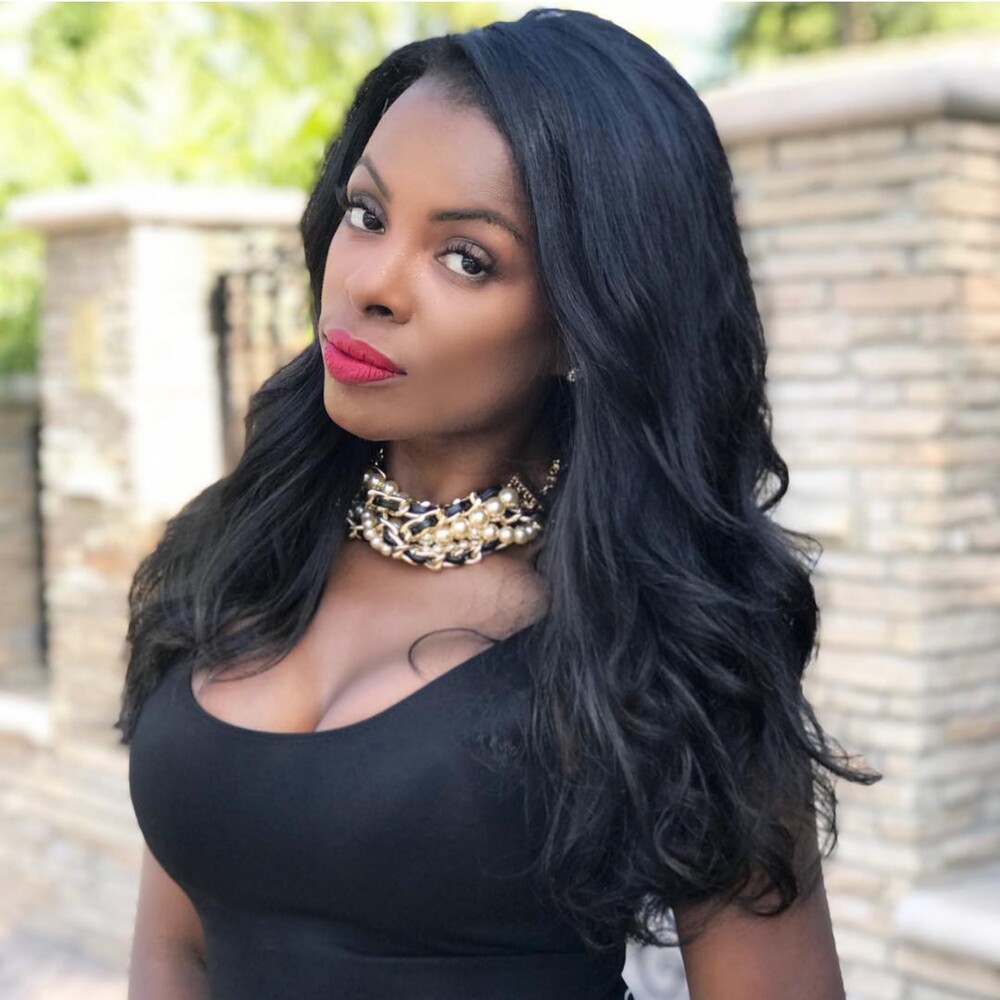 Eboni Berry with jet-black hair in loose waves past her shoulders, standing outside against a pale stone wall
