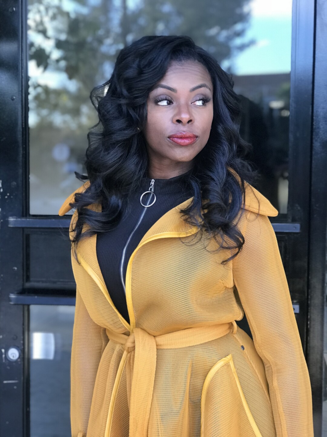 Eboni Berry looking to one side, her jet-black hair in loose curls past her shoulders, in a mustard mesh trench coat by a dark glass door