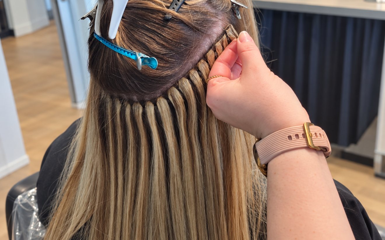 A hand pinching one bead on a row of no-sew beaded wefts, in dark-rooted balayage hair